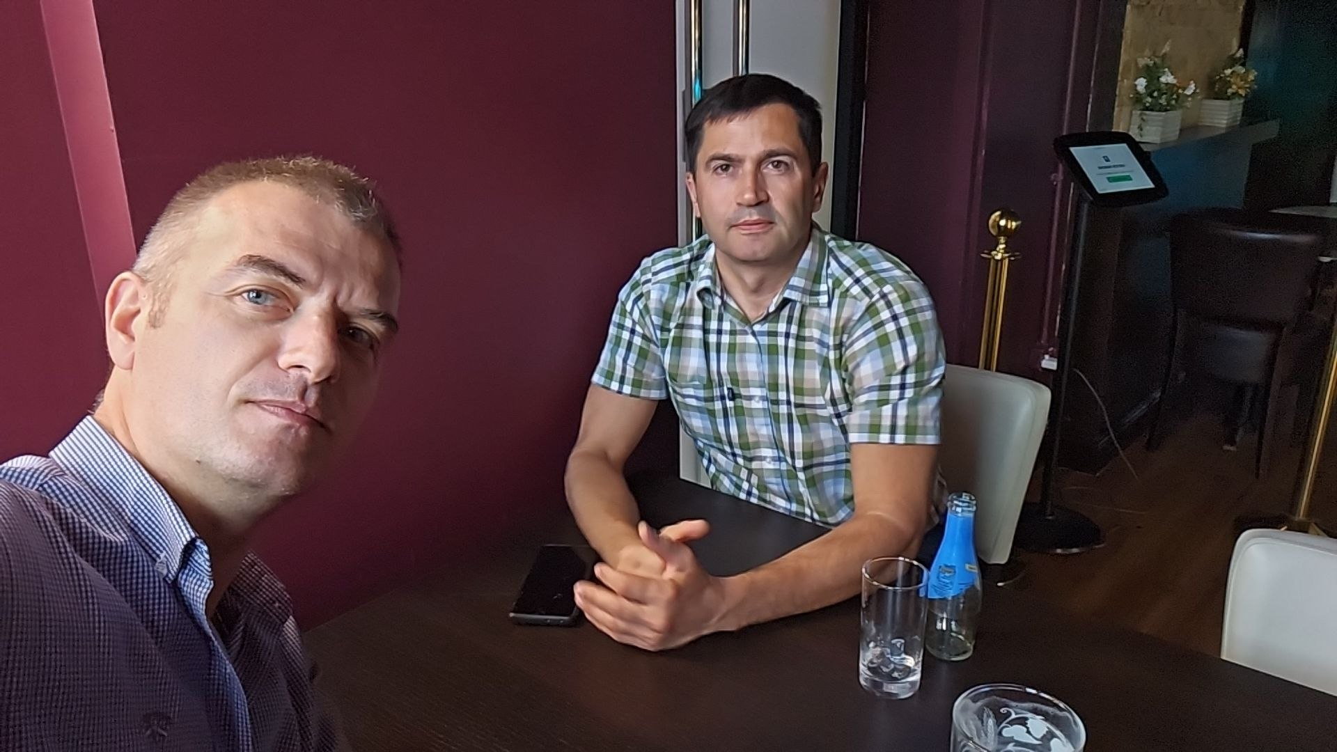 Two men seated at a dark wood table against a wine-red wall. The man on the right, in a green plaid shirt, sits with hands clasped. The man on the left holds the camera.